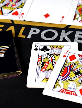 Poker-themed setup featuring a deck of cards on a table with the words 'REAL POKER' visible. Two cards, the King of Clubs and Jack of Diamonds, are prominently displayed. The design is vibrant, with detailed illustrations on the cards.