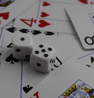 A collection of playing cards spread out, featuring various suits such as hearts, clubs, and spades. Two white dice with black dots are placed prominently on top of the cards.